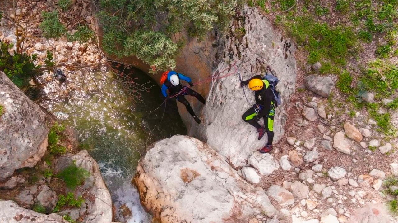 Two canyoners rappelling and managing an anchor station above a plunge pool